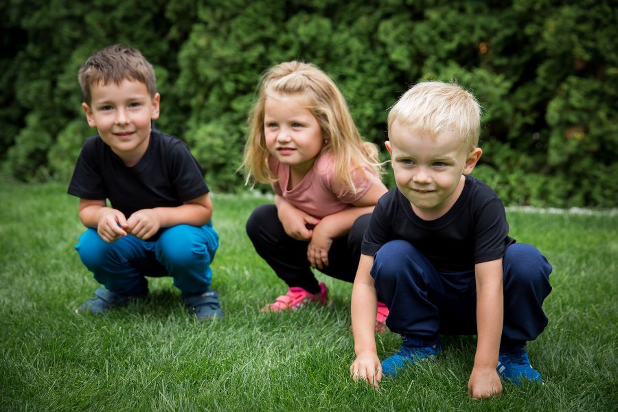 Tři děti sedí na bobku na trávníku, mají trička s krátkým rukávem a dlouhé kalhoty